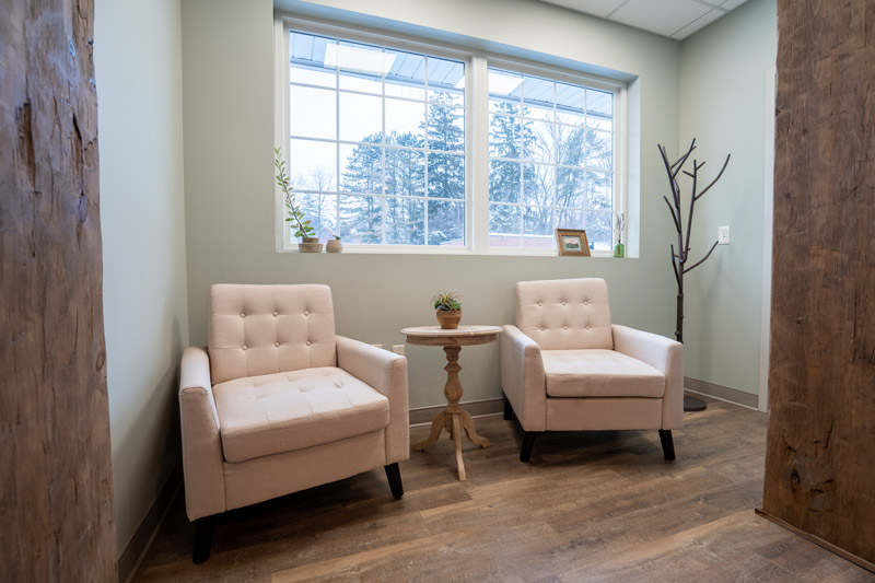 Cozy waiting area with two beige armchairs, a small wooden round table with a plant, large windows providing natural light, and minimal decor including a coat rack and potted plants.